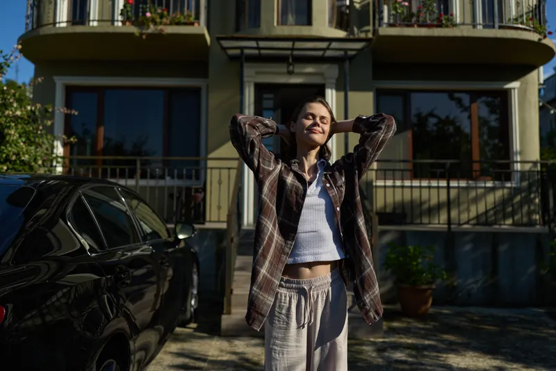 A woman in casual summer wear stands outdoors in front of a residential building, hands behind head, smiling, conveying relaxed lifestyle and everyday confidence
