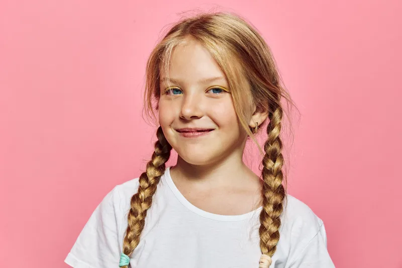 Smiling young girl with blonde braided hair and white shirt posing on a pink background studio portrait.