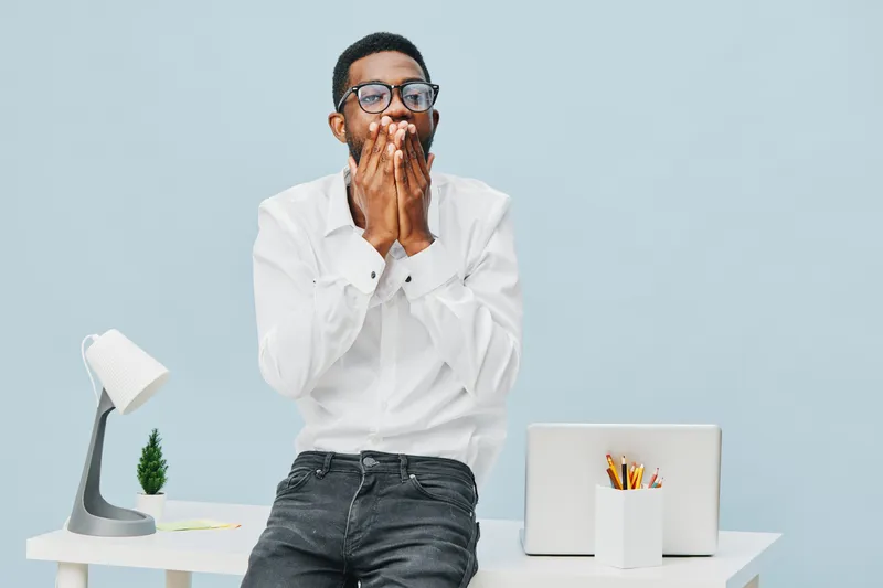 Modern expressive man wearing glasses and white shirt reacting surprised or shocked while sitting at a desk with laptop, stationery, and a small plant, isolated on plain light blue background. People 