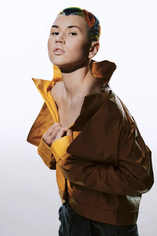 woman with colorful short hair wearing brown jacket posing confidently against a white background, stylish and modern portrait with artistic lighting effects