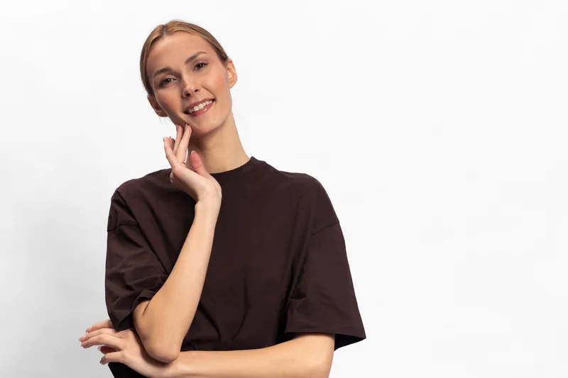 Smiling young woman in casual dark t shirt poses against white background with hand on face. Concept of positive emotions, confidence, youth, and natural beauty in studio portrait.