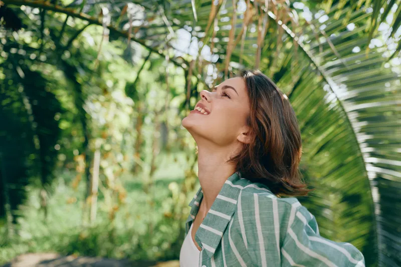 woman smiles outdoors in a vibrant garden setting, a joyful moment captured as she tilts her head back to feel the breeze, sunlight warming her expression.