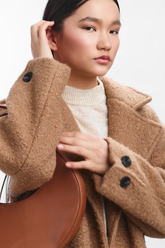 asian woman in a tan wool coat stands against a clean backdrop, adjusting her coat and holding a brown bag, radiating calm confidence and modern style.