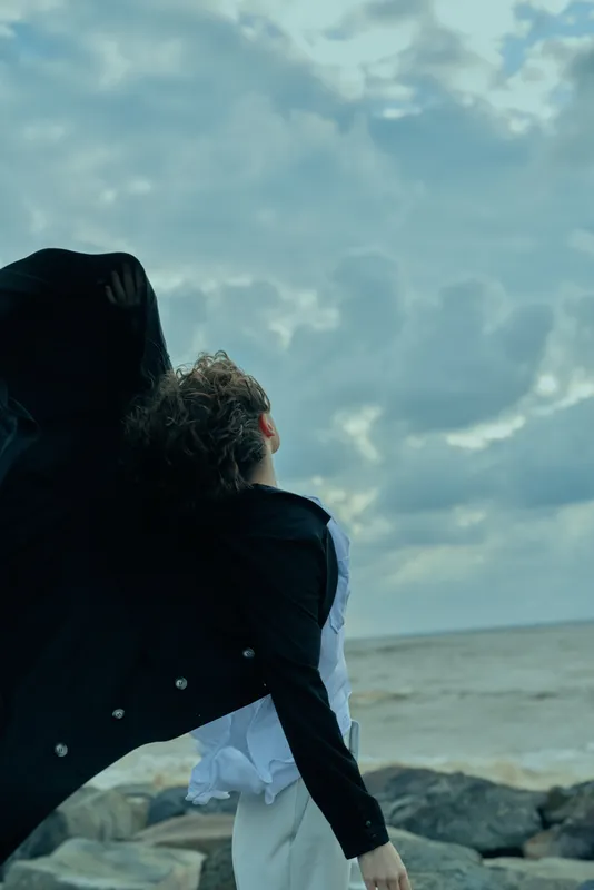 Couple embrace by the sea as a flowing coat billows in the wind, capturing a moment of connection, movement and hope on a rocky coast under a cloudy sky