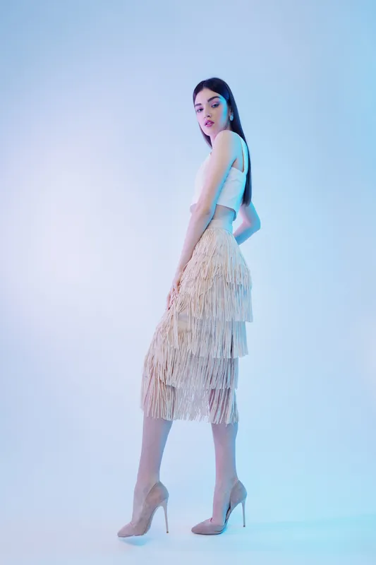 Elegant woman in a beige fringe skirt and white top poses confidently in high heels on a light background. Concept of beauty, fashion, and style.