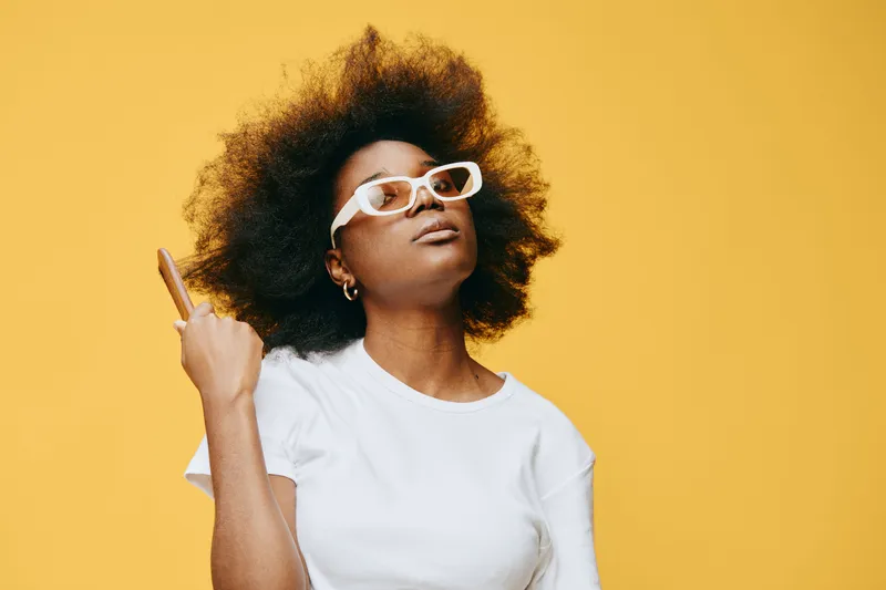 Confident black woman with curly hair in white glasses pointing up against a vibrant yellow background, showcasing empowerment and self expression in fashion
