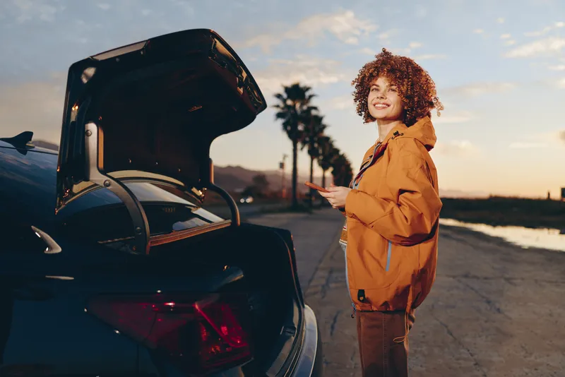 Woman lifestyle outdoors near car trunk at sunset, smiling with curly hair, wearing warm jacket, standing beside luggage, relaxed travel preparation, soft golden light, road scenery, moment