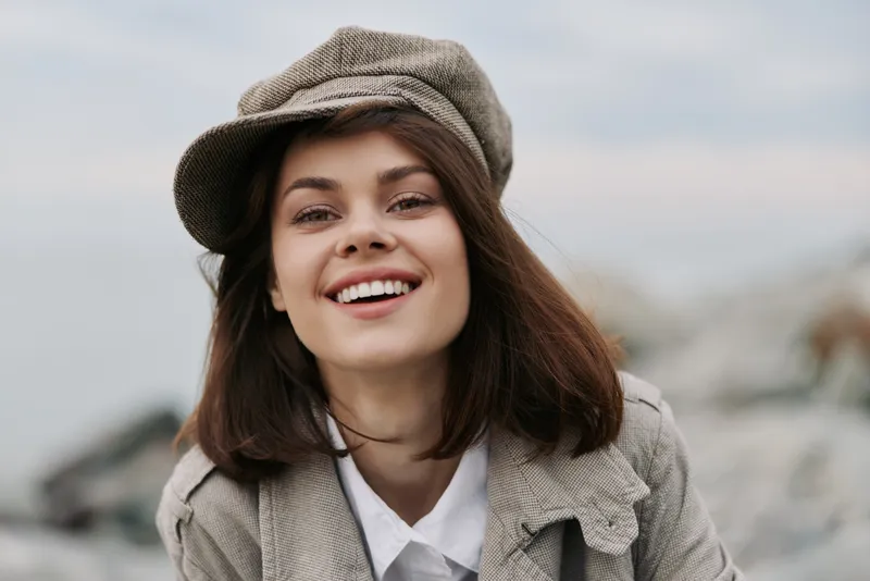 Smiling young woman wearing a textured hat and coat looks directly at camera outdoors. Natural light highlights her joyful expression and casual autumn style. Concept of happiness and fashion.