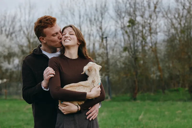 A smiling couple hug outdoors, a small dog cradled between them in a sunlit park. A warm, candid lifestyle moment that highlights affection, companionship, and simple happiness in everyday life.