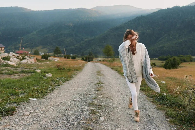 Woman outdoors, lifestyle scene on a mountain path, wearing a cardigan, white pants, boots, smiling with relaxed posture, sunlit breeze, natural colors, vast scenery