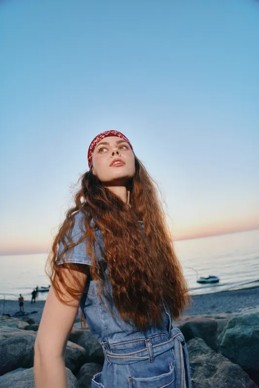 lifestyle woman with long hair wearing modern boho-western denim street style standing by the beach during sunset in film color