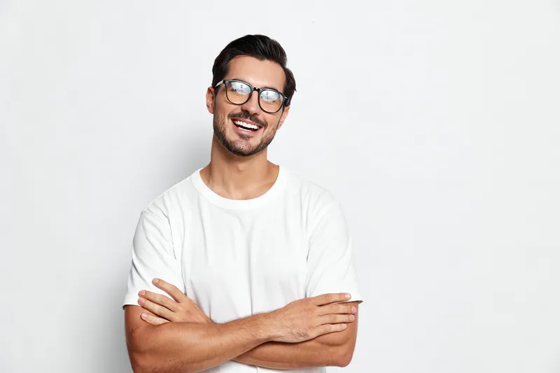 Smiling man with glasses standing confidently, arms crossed, wearing a casual white t-shirt, isolated on solid light background, showcasing a friendly and approachable demeanor.