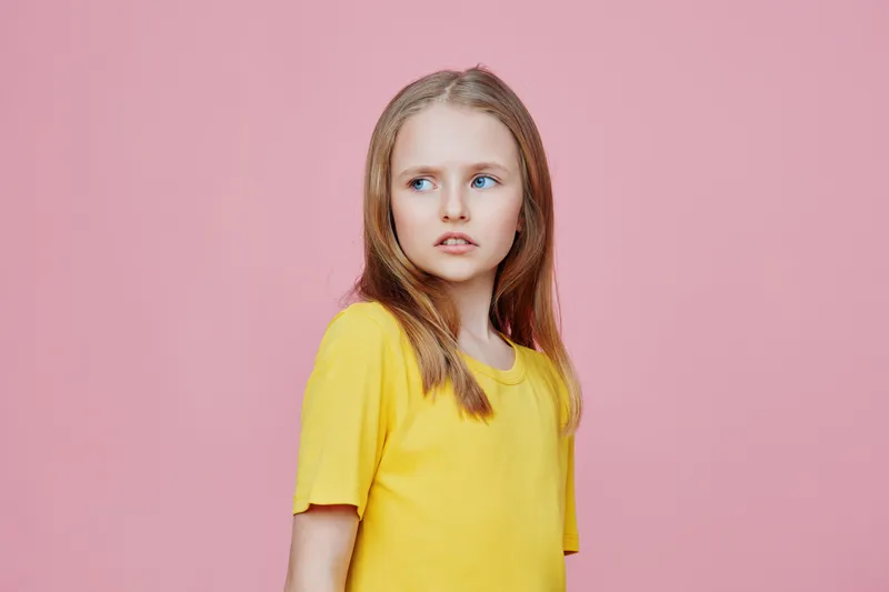 Young girl with long hair in a yellow shirt, looking thoughtfully against a pink background, capturing a sense of introspection and innocence