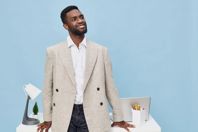 Confident smiling man standing near office desk with supplies, wearing stylish beige blazer over white shirt, implying professional success and positive attitude. Studio shot on plain blue background.