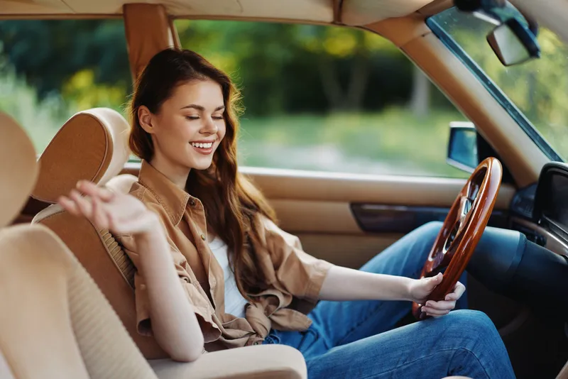 Young woman enjoying a drive in a vintage car, smiling and relaxed, wearing a casual outfit, set in a green outdoor environment during summer.