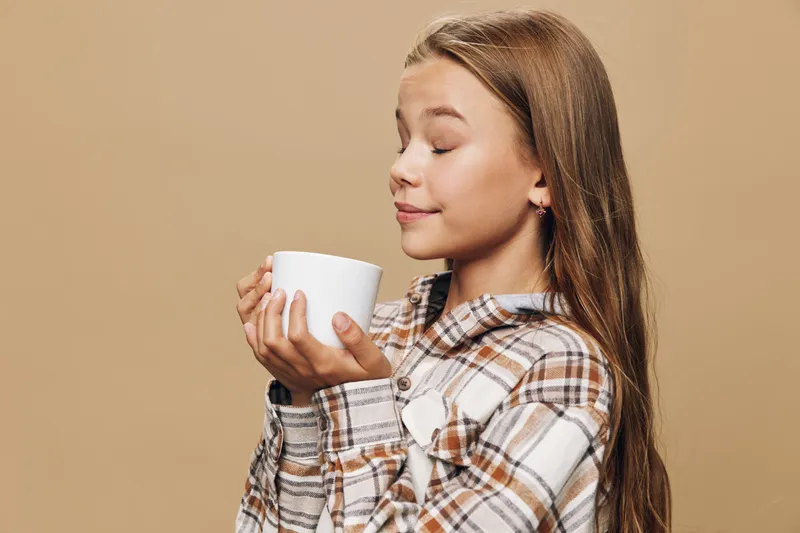 Girl with long hair holds white cup close to face eyes closed enjoying aroma with peaceful smile wearing checked shirt on beige background