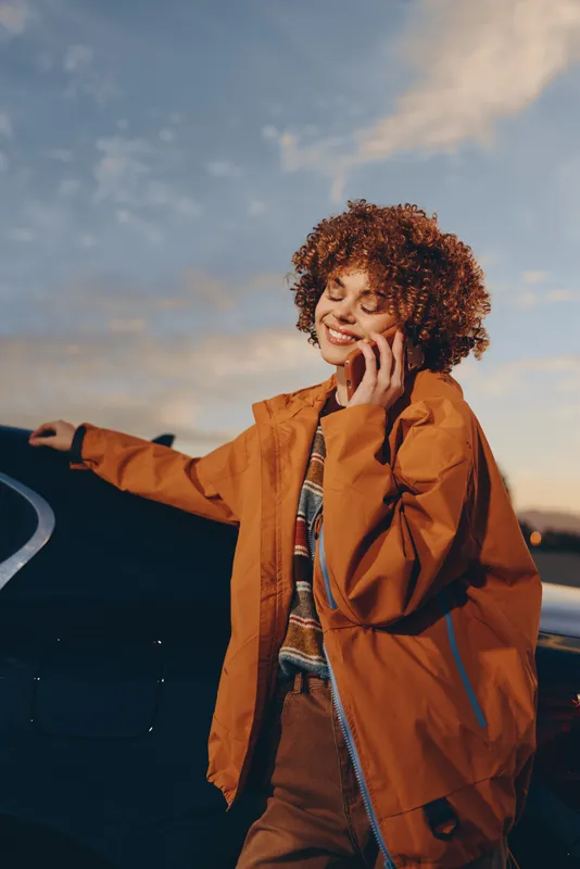 Woman with curly hair stands outdoors near a car at sunset, wearing a coat, one hand on her face, lifestyle portrait with travel mood, warm sky, calm fashion, and confidence