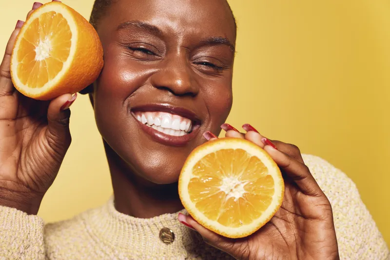 Smiling African woman with dark skin holds two orange halves near her face on yellow background. Concept of healthy lifestyle, vitamin C, skincare and natural beauty with fresh fruit.