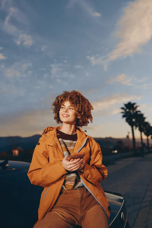 Woman lifestyle portrait outdoors sunset road curly hair smiling jacket brown, sitting near car on roadside, hands clasped at chest, warm evening light, urban travel mood, soft sky clouds