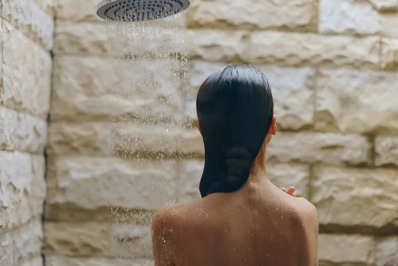 Woman taking outdoor shower with wet hair under rain showerhead, natural stone wall background, calm and refreshing moment.