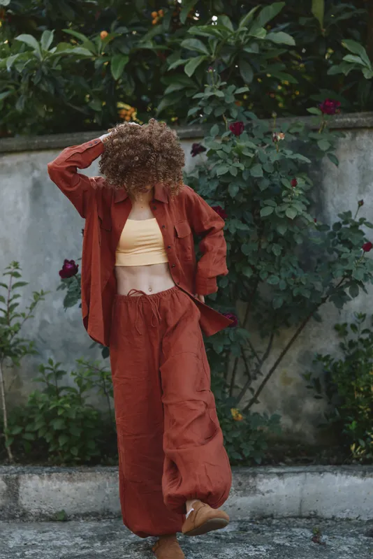 Young woman with curly hair in a rust-colored tracksuit dances playfully in a garden, radiating carefree style, relaxed mood, and warm outdoor light