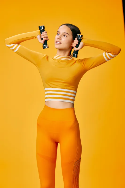 Fitness woman in an orange workout set performs an exercise with dumbbells up by the head, striking a confident pose in a bright orange studio backdrop.