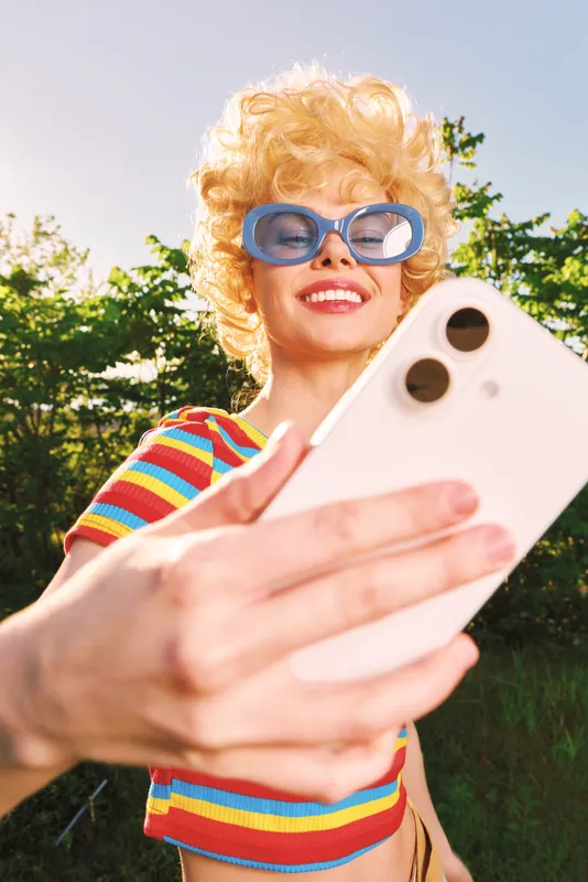 Selfie minded woman with curly hair wearing retro sunglasses outdoors under sunny light, colorful striped shirt, smiling while holding a smartphone, portraying casual lifestyle and joy.