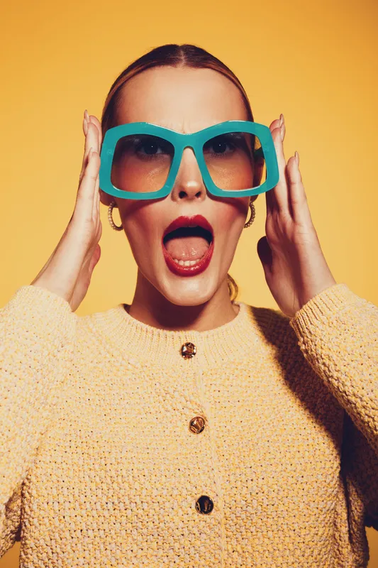 Surprised woman in oversized blue sunglasses and yellow sweater poses against yellow background. Fashion portrait with bold accessories and expressive facial expression.