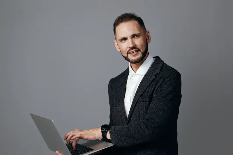 Professional businessman on a neutral colored background, displaying confidence and focus while working on a laptop, conveying dedication, leadership, and strong work ethics.
