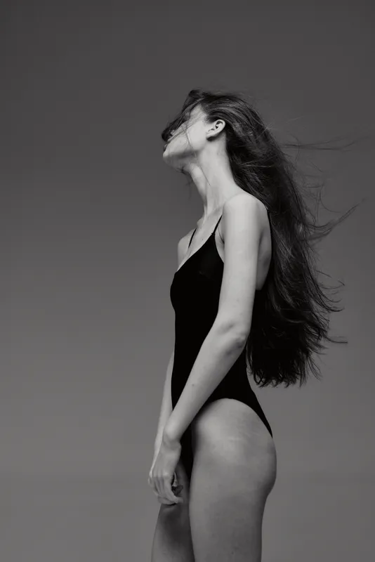 Black and white artistic portrait of a slender woman posing gracefully in a swimsuit with long flowing hair against a neutral background.