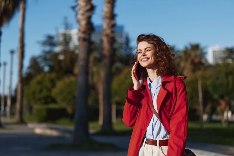 A young woman in a red coat smiles while talking on her mobile in a sunny park with palm trees, wearing a light blue shirt and beige trousers, creating a bright lifestyle moment.