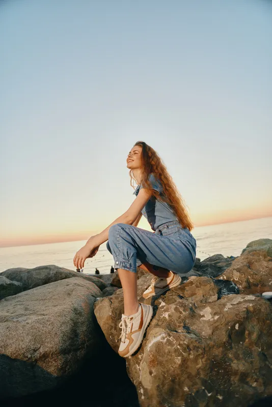 Lifestyle woman in modern boho-western denim street style sitting on rocks by the sea, captured with warm film color during sunset, relaxed and contemplative mood.