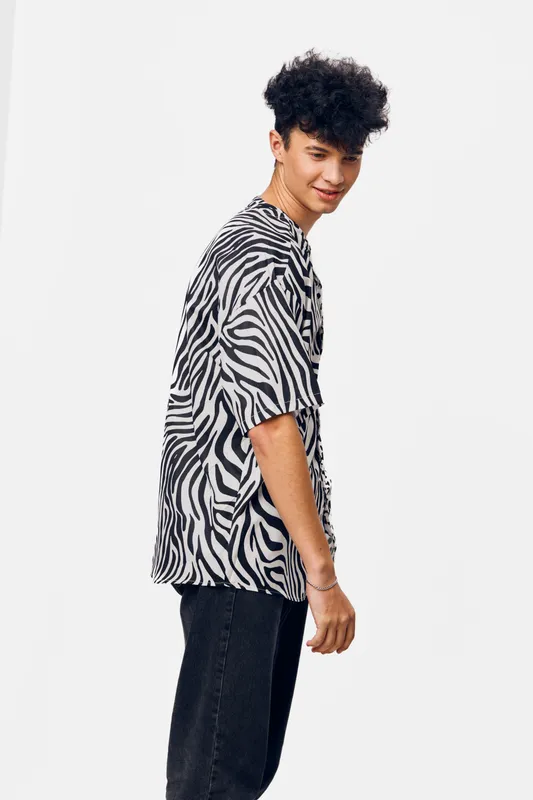 young man wearing a zebra print shirt standing sideways with a playful smile against a white background, casual style and curly hair