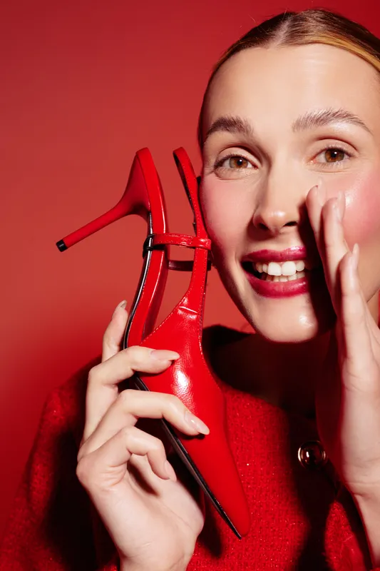 Smiling woman in red sweater holds red high heel shoe near face on red background. Concept of fashion, style, and vibrant color coordination with joyful expression and modern look.