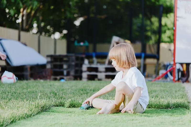 A thoughtful boy playing with a toy car on a sunny day in a green backyard, enjoying outdoor activities with toys and friends, surrounded by nature.