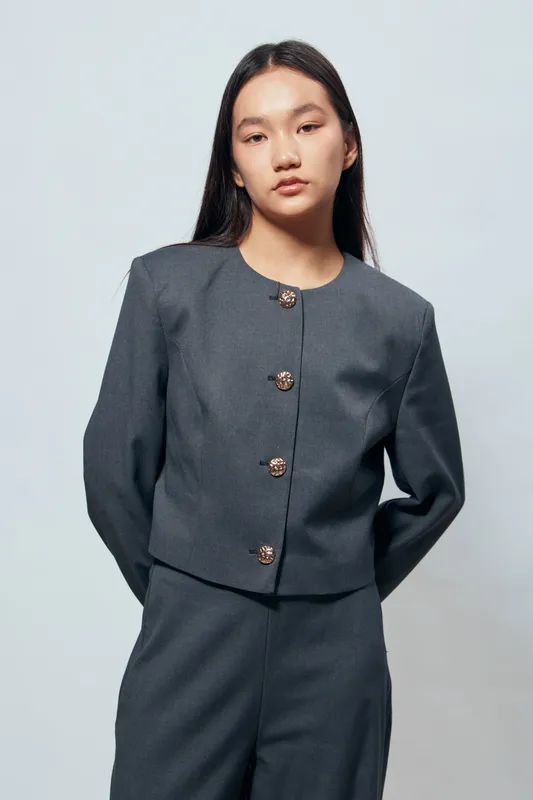 Young woman portrait in gray suit jacket and pants standing with hands behind back on light background. Fashion model with long dark hair in simple studio setting looking at camera.