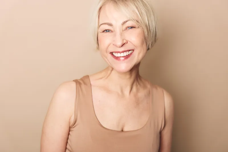 Smiling mature woman with short blonde hair wears beige tank top against neutral background. Natural beauty concept with positive emotions and healthy skin in warm soft lighting portrait.