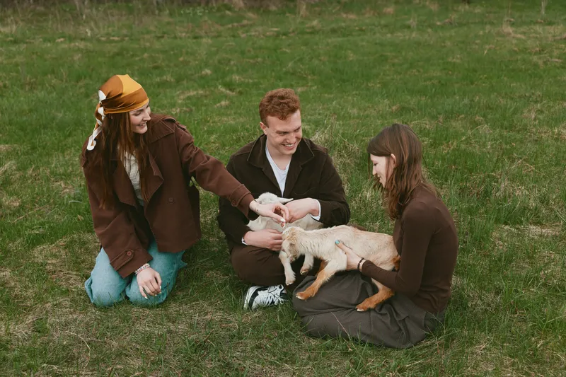Group of friends enjoys an outdoor moment with a friendly goat kid, pets it gently, shares smiles, casual lifestyle scene on a grassy field, warm tones, candid interaction and curiosity.