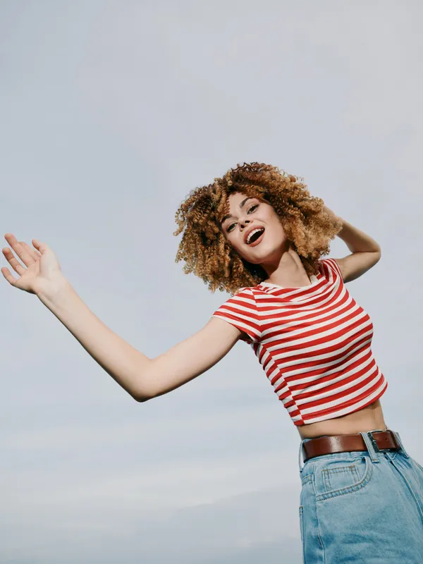 A joyful young woman with curly hair wears a red and white striped top and blue jeans, arms outstretched in a carefree pose, smiling beneath a bright blue sky.