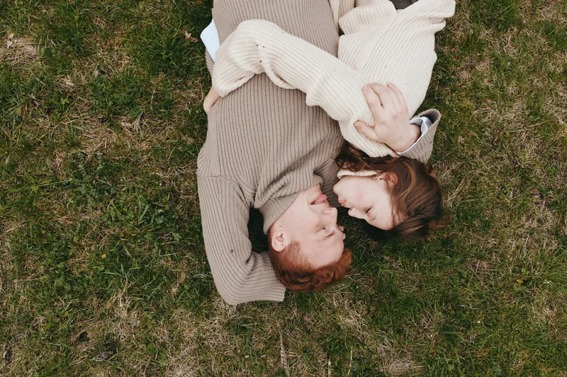 Relaxed couple lying on grass, embracing softly in a warm outdoor lifestyle moment, candid and intimate, natural light, cozy sweaters, peaceful mood, romantic countryside vibe and serene air
