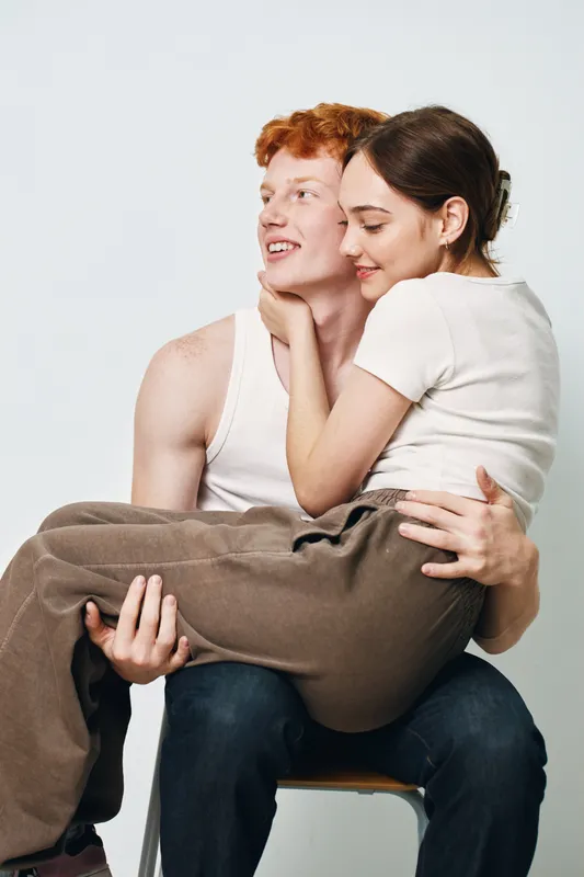 Couple sitting and embracing with happy expressions, man holding woman on lap in casual clothes. Portrait of young people in relaxed pose with affection and close connection.