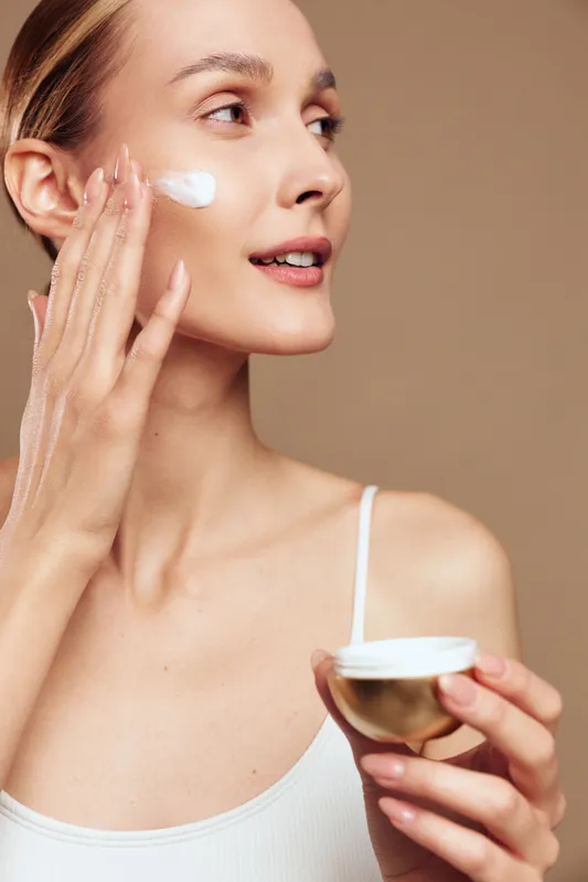 Woman applying facial cream and moisturizer to cheek, holding a small bowl, calm expression, spa beauty routine, self care concept, studio close up on beige background