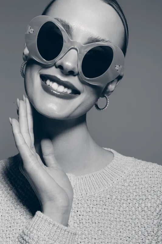 Smiling young caucasian woman in round sunglasses and knitted sweater poses with hand near face on grey background. Fashion and beauty concept.