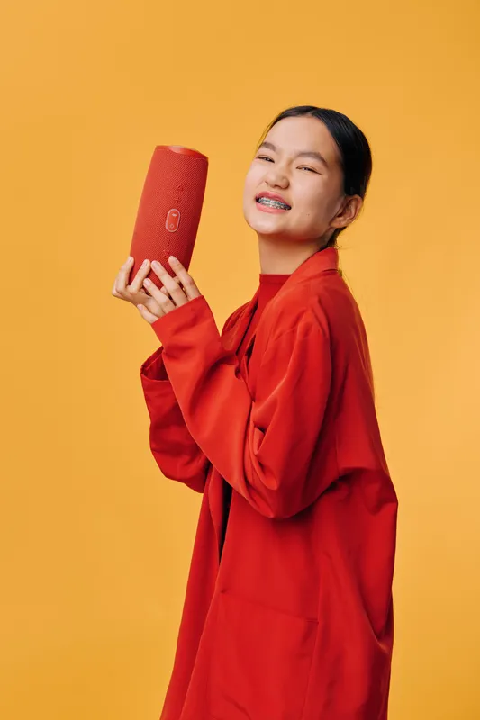 Young woman wearing braces and red outfit holding red textured clutch bag smiling with eyes closed against yellow background. Fashion accessory and trendy style with casual look.
