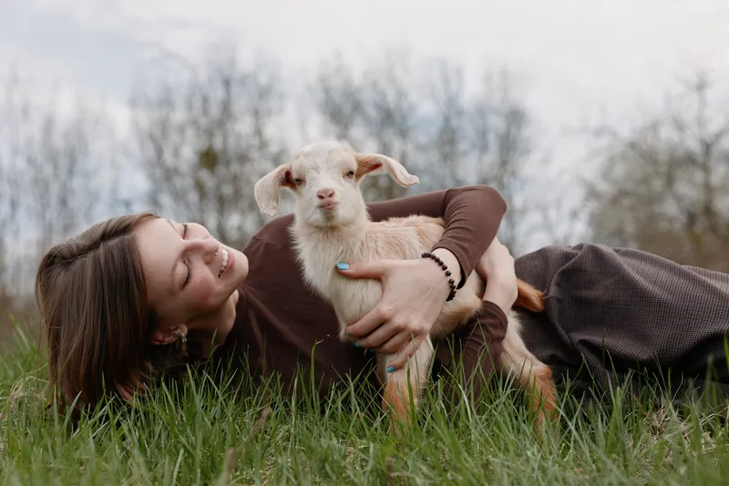 Young woman lying on grass embraces a baby goat outdoors in a natural setting, smiling gently and enjoying a peaceful lifestyle moment in casual clothing.
