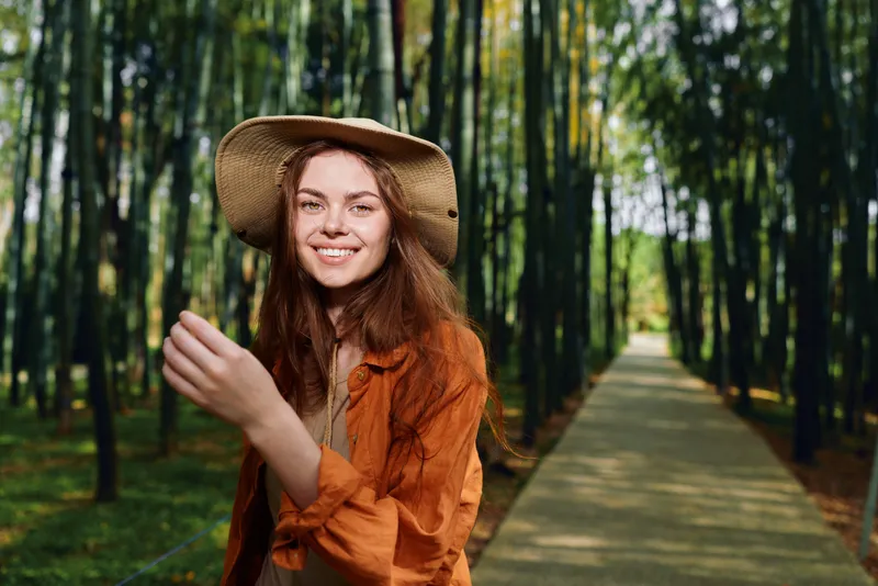 Woman smile hat forest path walkway nature portrait of a happy young female outdoors on a wooden boardwalk, candid travel lifestyle photo with sunlight and relaxed expression