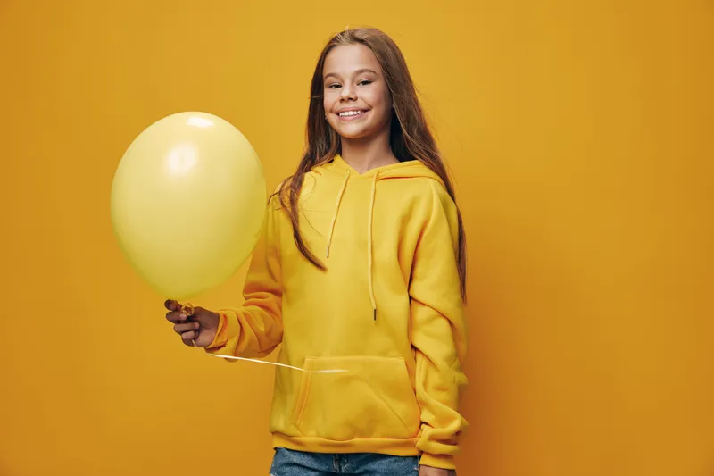 Girl with smile holding yellow balloon wearing yellow hoodie and denim jeans on yellow background. Portrait of happy child with long hair enjoying party or celebration.