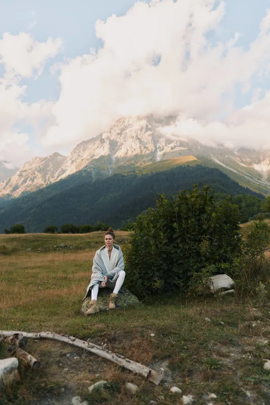 A solitary man sits on a rock beside a bush in a mountain meadow, wearing a light jacket and jeans, enjoying the open air and tranquil scenery.