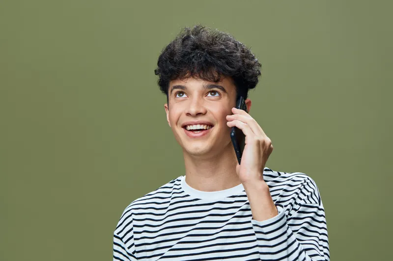 smiling young man in striped shirt talking on mobile phone with cheerful expression against green background, communication concept, casual style, modern technology