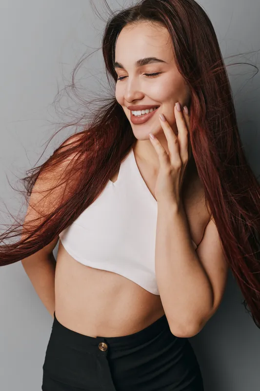 Young woman with long hair smiling with eyes closed wearing white crop top and black pants posing against gray background. Beauty, happiness and casual style in studio portrait.